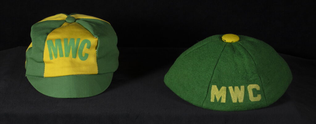 Two caps sit side by side on a dark surface, one with alternating green and yellow panels and the other solid green, both displaying the letters “MWC” on the front.