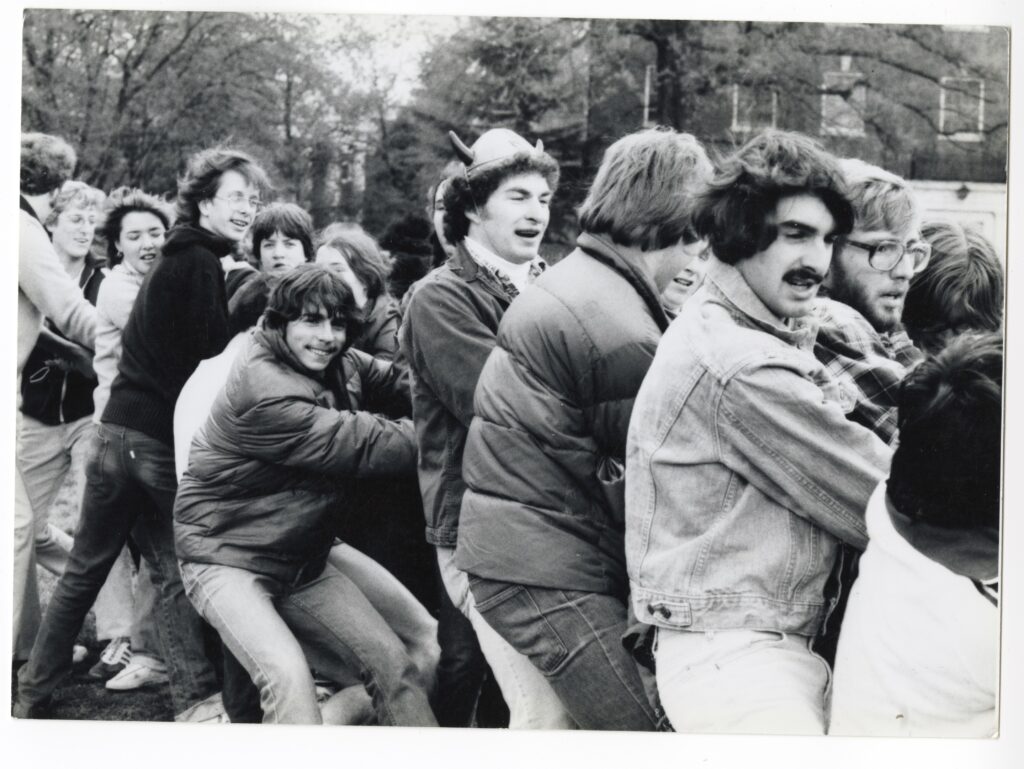 A group of people outdoors pulls together on a rope, standing close in cool‑weather clothing, and some with devil horn hats, with trees and buildings in the background.