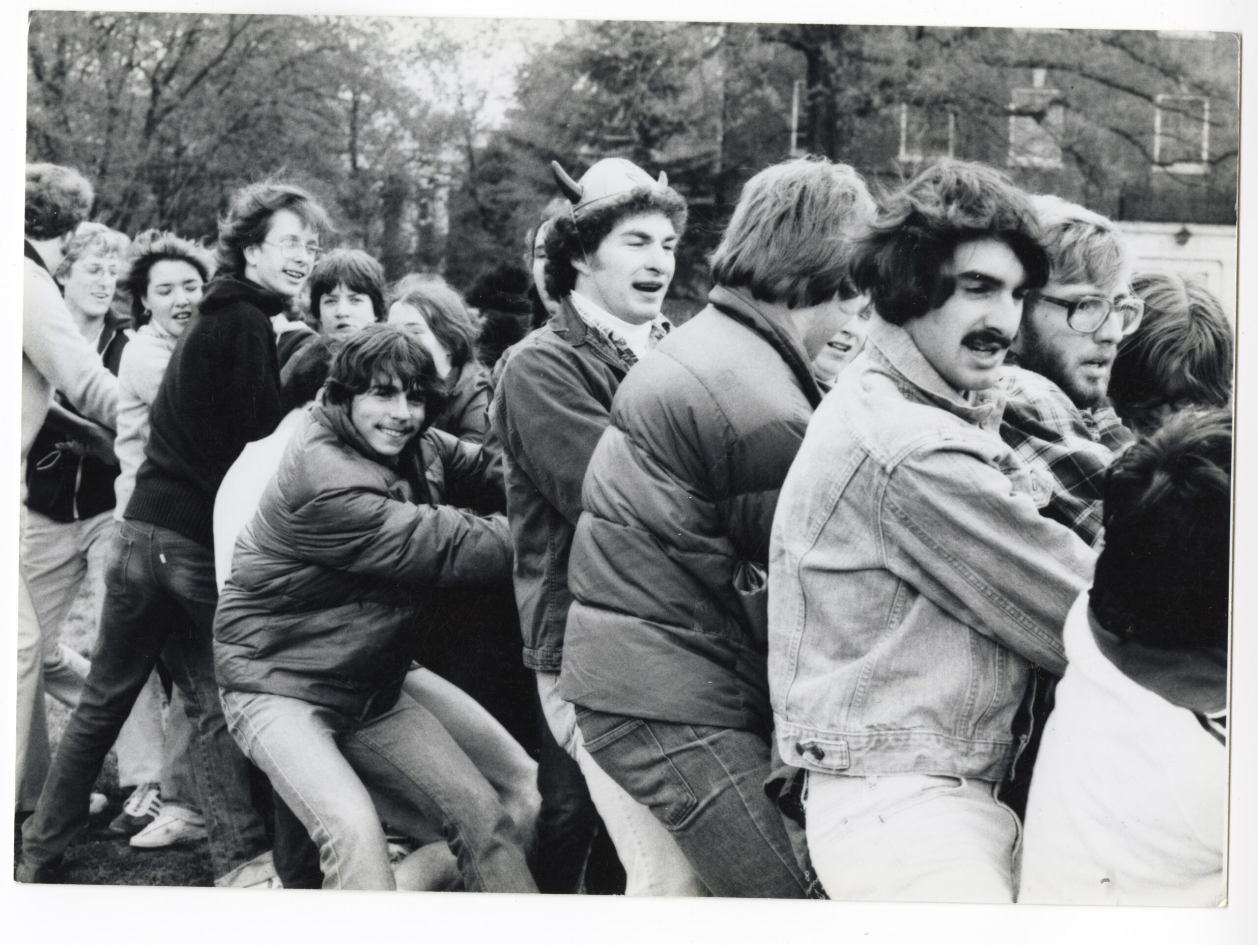 A group of people outdoors pulls together on a rope, standing close in cool‑weather clothing, and some with devil horn hats, with trees and buildings in the background.