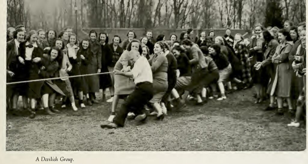 A large group of people outdoors pulling on a rope in a tug‑of‑war, with spectators gathered around on a grassy field bordered by leafless trees and the caption “A Devilish Group.”