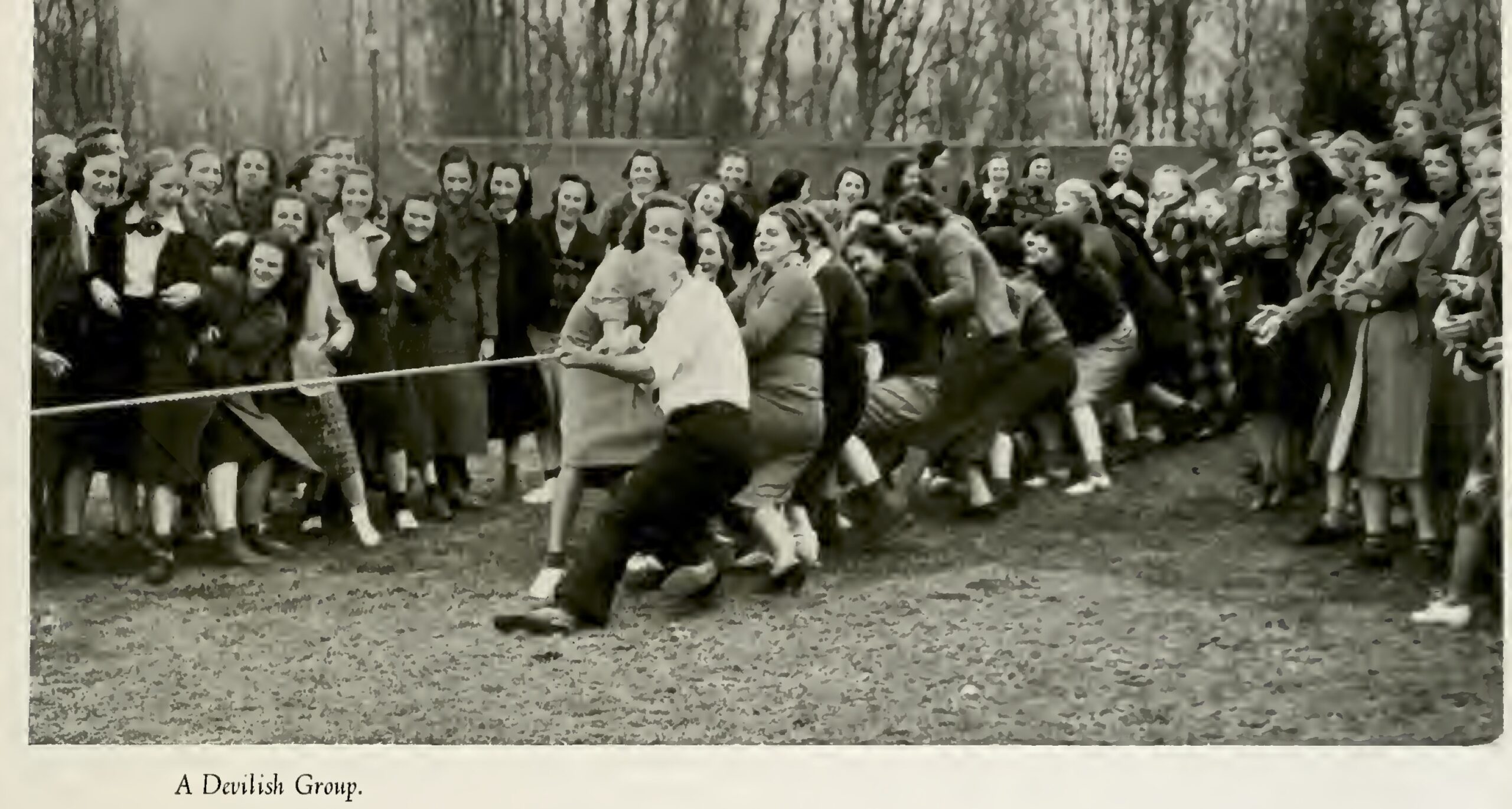 A large group of people outdoors pulling on a rope in a tug‑of‑war, with spectators gathered around on a grassy field bordered by leafless trees and the caption “A Devilish Group.”