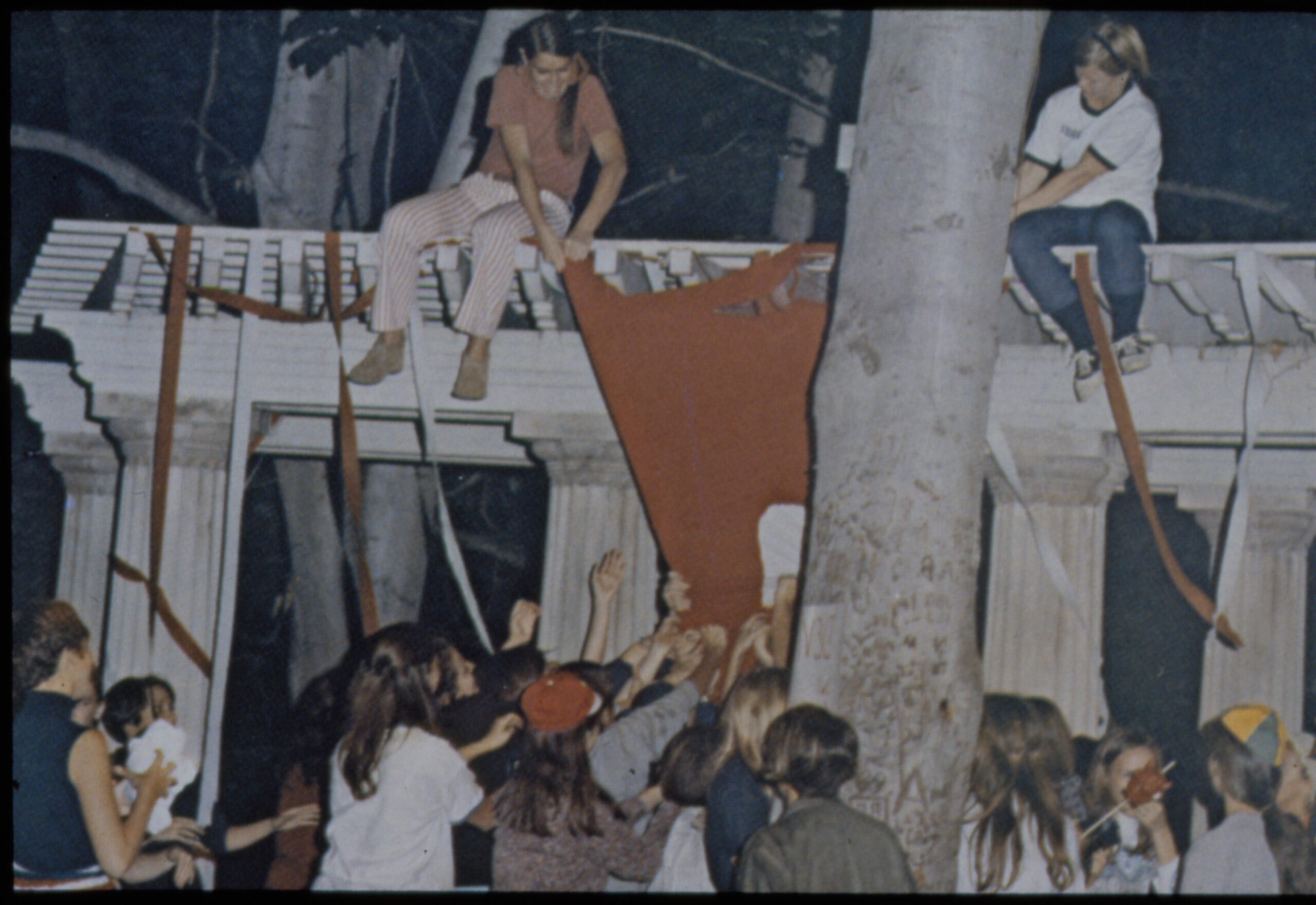 People climb onto a columned structure at night and hang long red and orange fabric strips down to a crowd reaching up toward them.