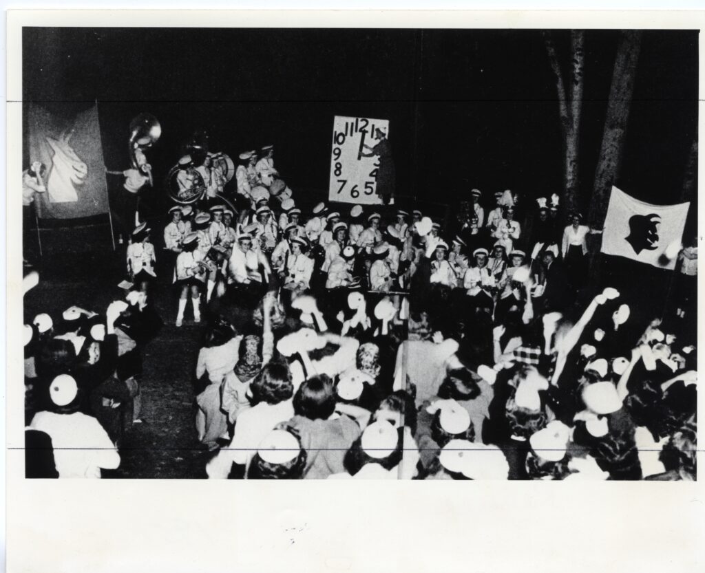 Dozens of students and band members surround a large countdown clock with a banner with a devil and a separate banner with a goat, which are on the left and right.