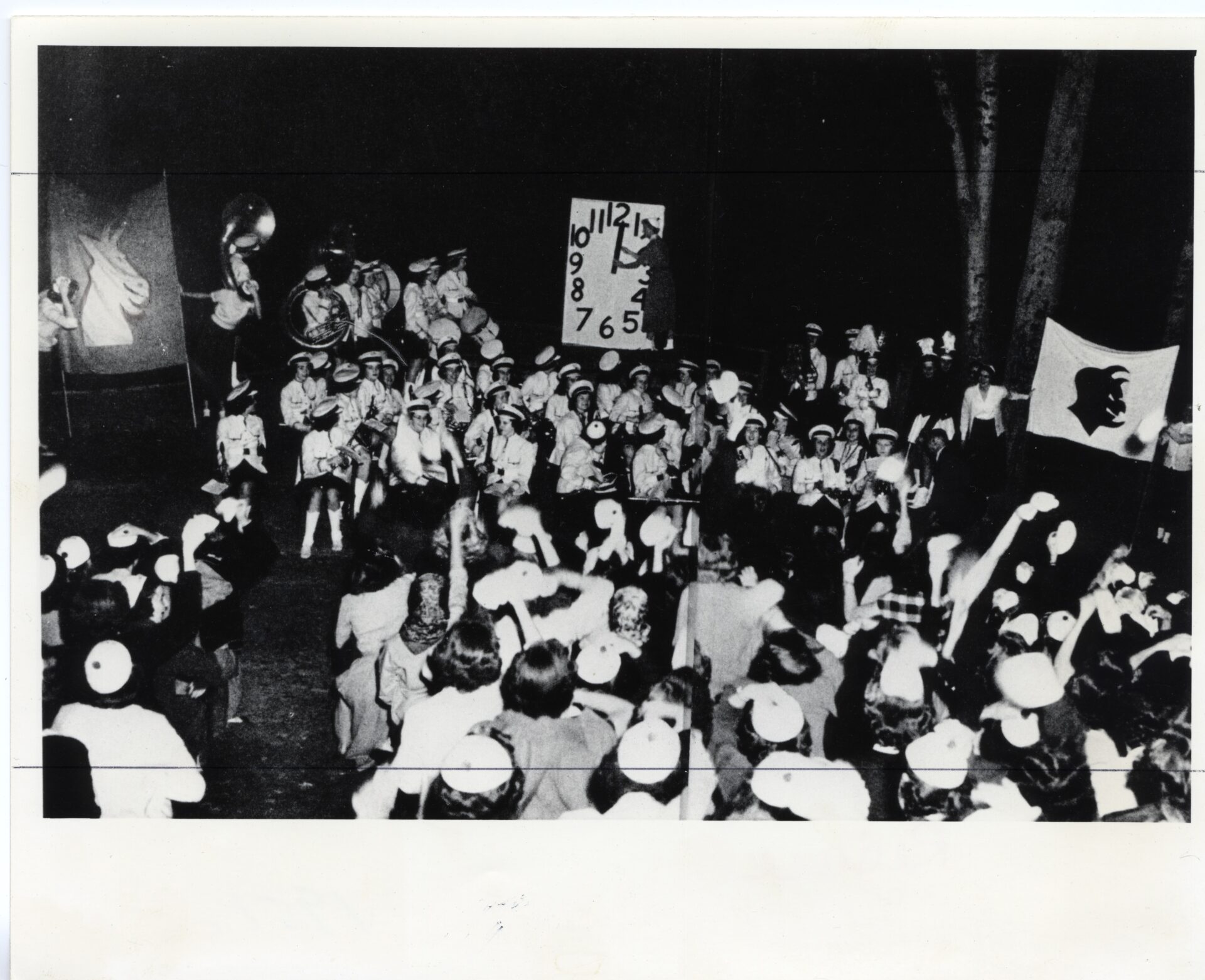Dozens of students and band members surround a large countdown clock with a banner with a devil and a separate banner with a goat, which are on the left and right.