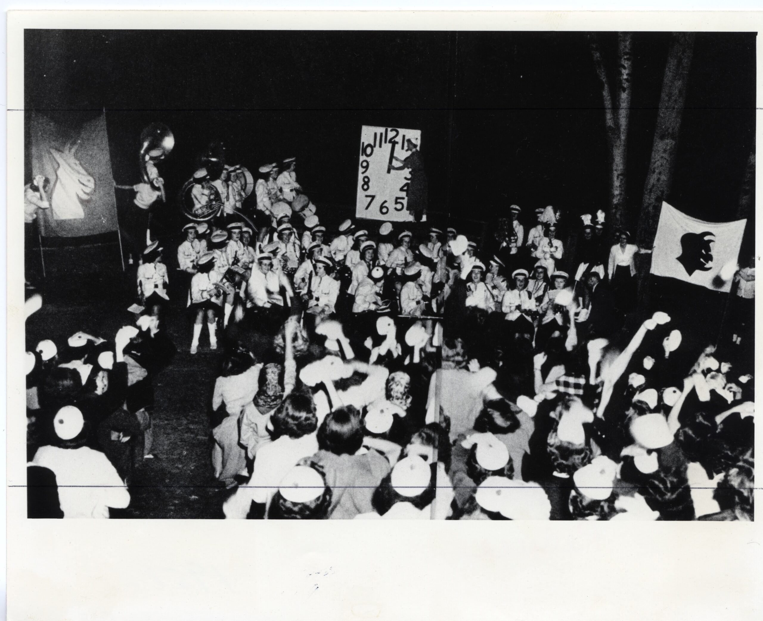 Dozens of students and band members surround a large countdown clock with a banner with a devil and a separate banner with a goat, which are on the left and right.